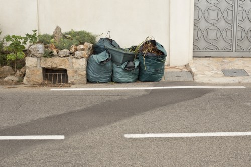Four large, dark green garbage bags filled with waste are placed on the pavement next to a cream-coloured building with a small, rectangular subterranean vent at ground level. The bags appear to contain mixed rubbish, possibly household or garden waste, and are positioned adjacent to a small patch of overgrown plants and rocks that are part of a low, weathered stone wall. The street surface in the foreground is asphalt with visible white road markings, indicating a quiet urban or residential area potentially within or near Bayswater, London. To the right side of the image, a metal door with an intricate, decorative pattern is partially visible, mounted on the building's wall. The scene illustrates typical rubbish accumulation awaiting collection or disposal, and it is managed by a professional waste removal service such as Office Clearance Bayswater, serving local residents and businesses.