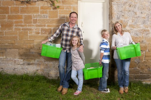 A family of four standing outdoors against a weathered brick wall near a white door, each holding bright green plastic storage boxes. The father, positioned on the left, is wearing a plaid shirt with rolled-up sleeves and jeans, smiling and holding one box in each hand. The daughter, standing close to her father, is wearing a grey patterned dress and has long blonde hair, holding a box in front of her. To their right, a young boy in a blue and white striped shirt and jeans is holding a green box and looking towards the camera. Further right, the mother, dressed in a white blouse and jeans, is also holding a green storage box and standing beside her son. The scene suggests they are preparing for rubbish removal or clearance, possibly in the Bayswater area, with the family standing on a patch of grass and the setting implying a residential environment that could relate to domestic clearance services offered by Office Clearance Bayswater in London postcode W2.