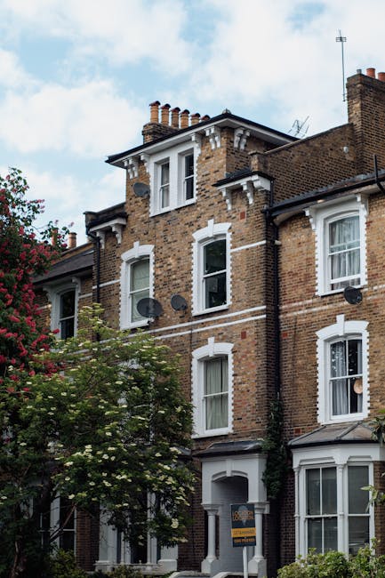 A multi-storey Victorian-style brick residential building with a traditional dark brown brick facade and white window frames situated on a street with a partly cloudy sky above. The building has three