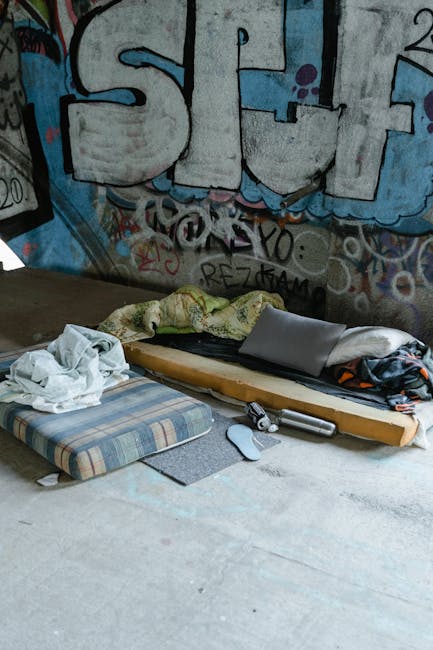 A minimalist bedroom featuring plain white walls and a wooden floor in a neutral tone. The room contains a double bed with a simple wooden headboard and black metal legs, positioned centrally against 