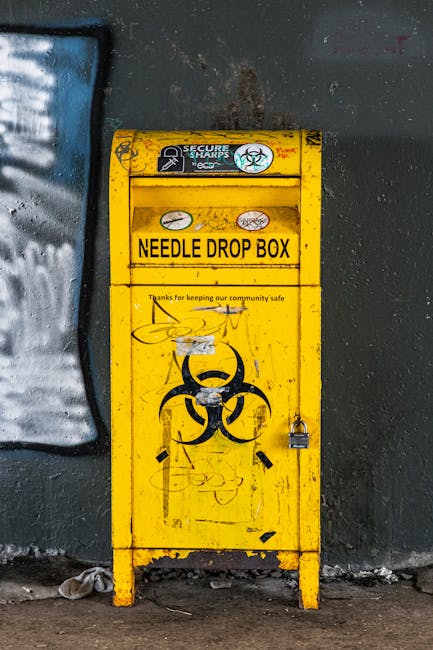 A tall, narrow yellow needle drop box made of metal, positioned against a dark, textured wall with areas of peeling paint and graffiti. The front surface features a black biohazard symbol and some gra